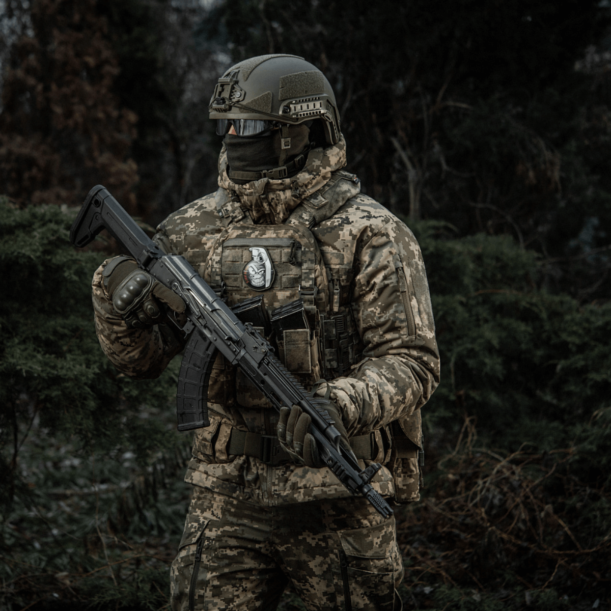 Soldat i camouflage med riffel i skov, bærer M-Tac Patch M67 Granat 3D PVC på taktisk vest.
